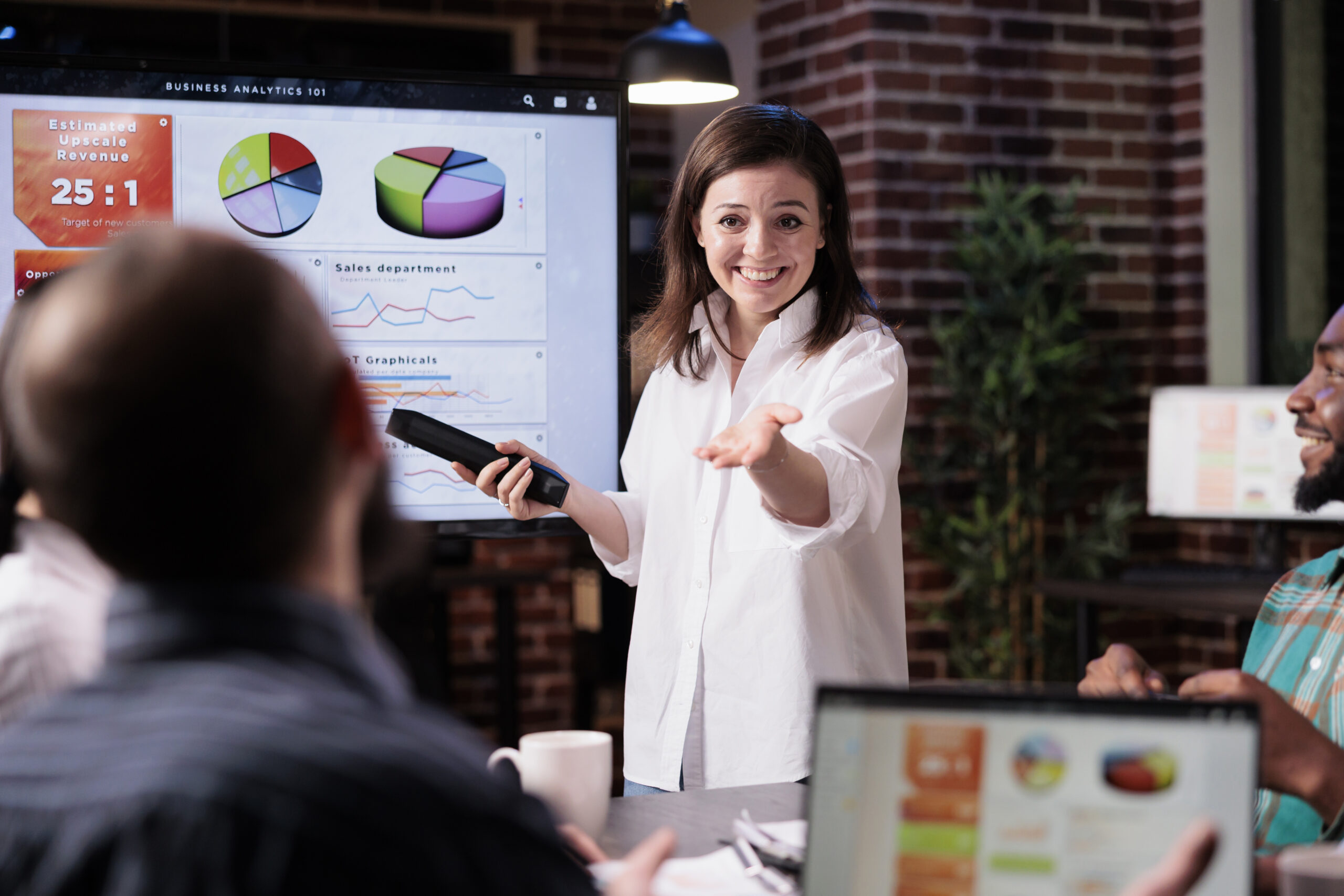 Smiling business employee standing in front of tv screen with sales statistics talking with man with laptop